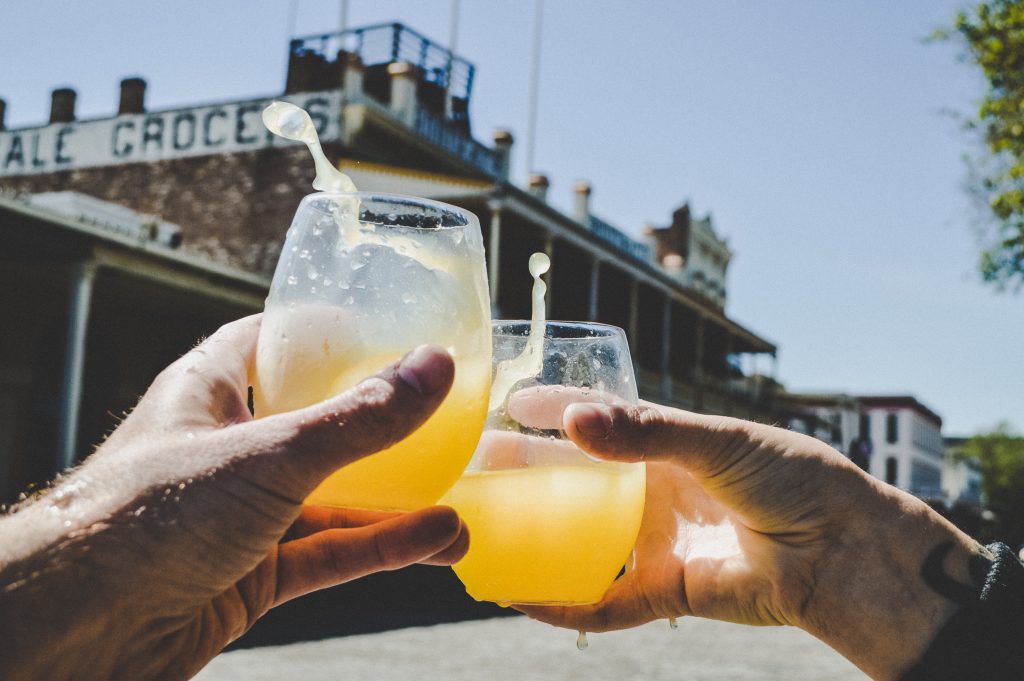 Two hands raise glasses filled with orange drinks in a celebratory toast, liquid splashing. A historic brick building is visible in the sunny background.