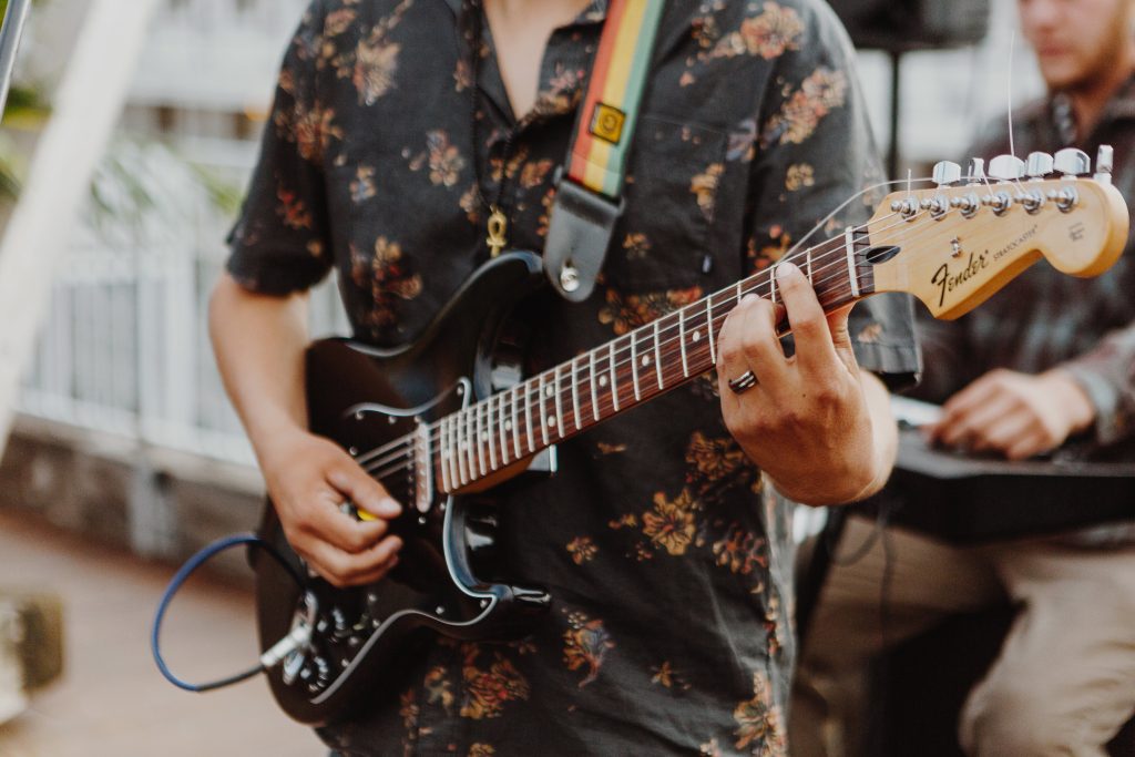 A person in a floral shirt plays an electric guitar with a rainbow strap. Another person plays a keyboard in the blurred background, creating a lively atmosphere.