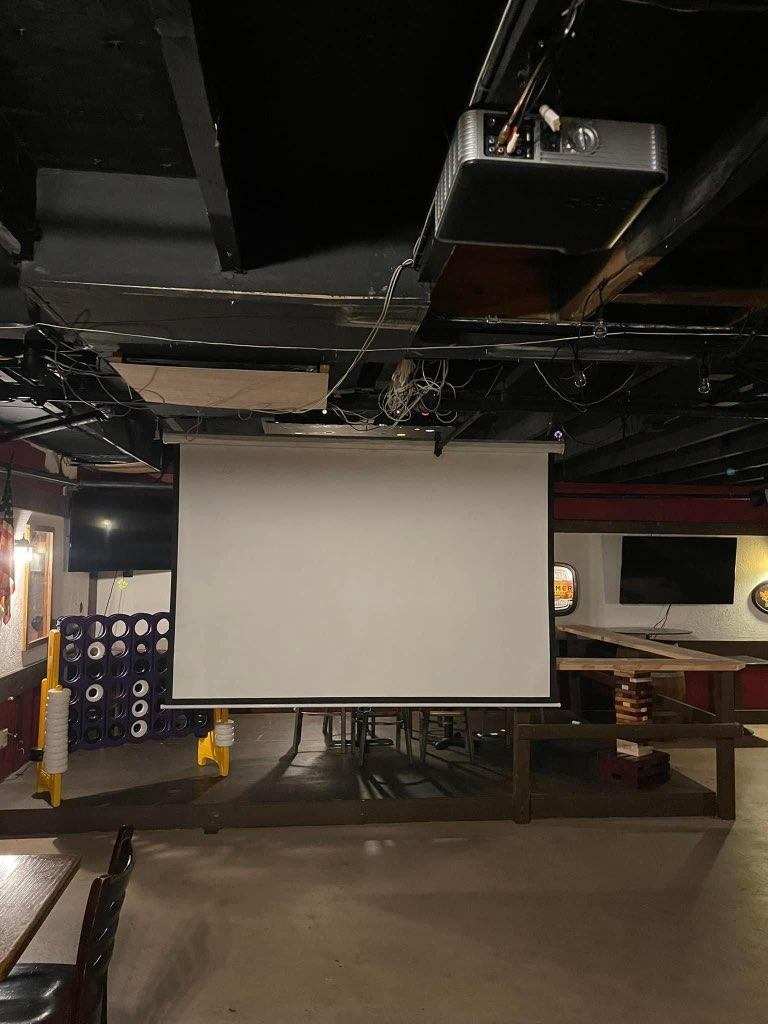 Pull-down projector screen hanging from the ceiling over an open floor area at O’Mally’s, with a ceiling-mounted projector and wall TVs