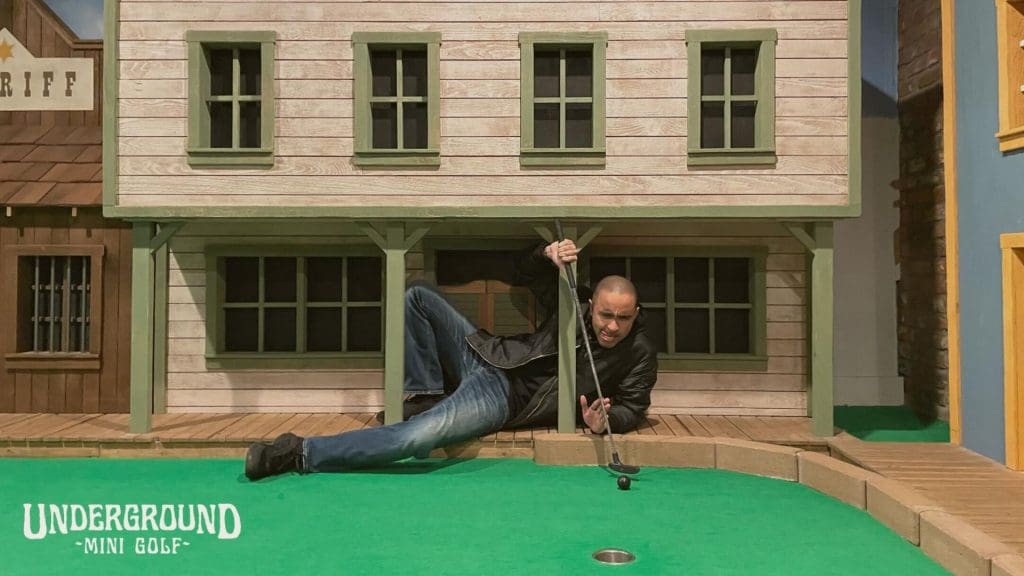 Man lying on his side on the putting green at Underground Mini Golf, lining up a putt on a hole set in front of an Old West town facade