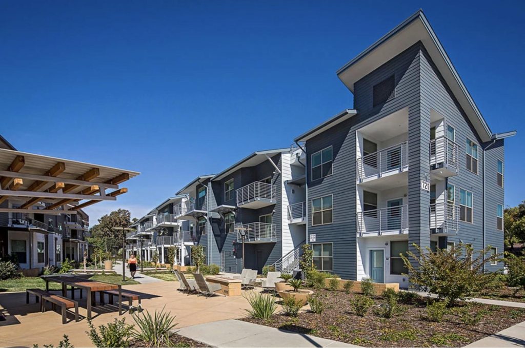 Modern apartment complex with blue-gray siding and angular roofs set against a clear blue sky. Features balconies, a shaded patio area, and minimal landscaping.
