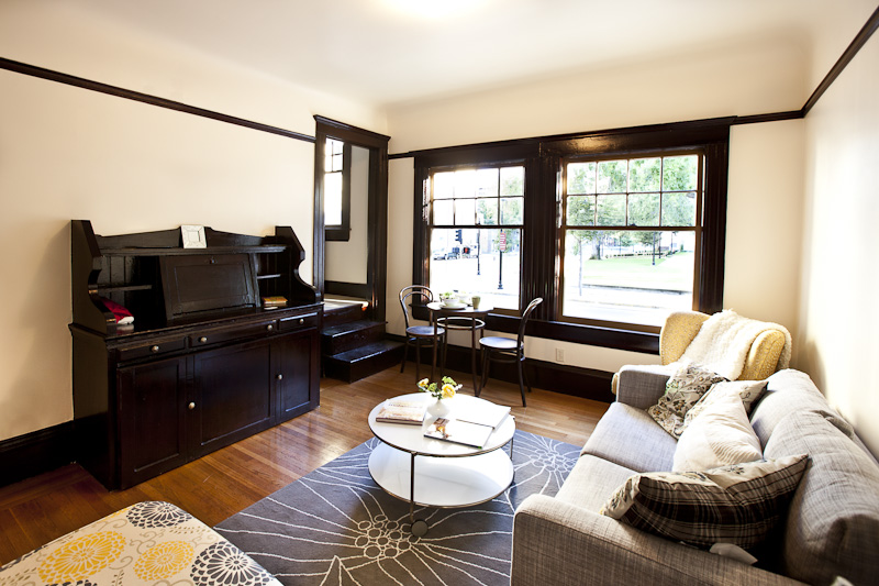 Cozy living room with a gray sofa, patterned rug, and round coffee table. Dark wood furniture and trim, large windows overlooking a park. Warm, inviting ambiance.