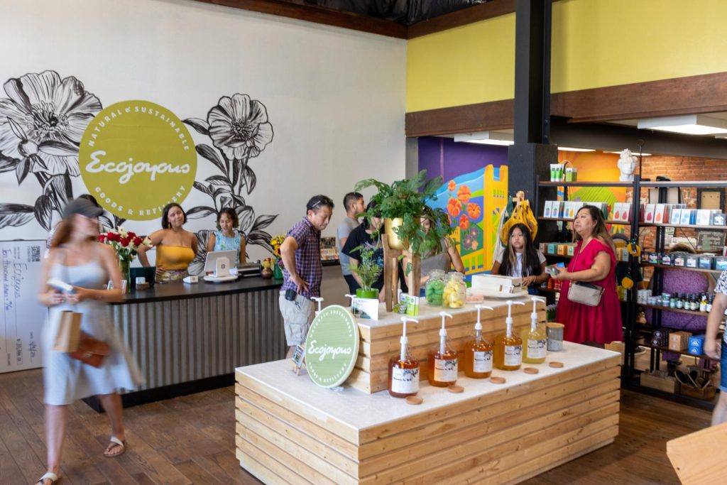 A bright, bustling eco-friendly store, with a display of natural products and plants. Customers browse near a floral mural and yellow walls. Friendly, vibrant atmosphere.