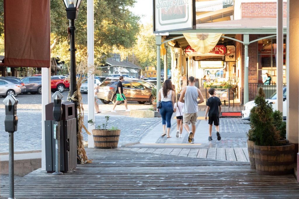 A family strolls down a charming wooden sidewalk in a bustling, sunlit town center. Trees, cars, and a vintage-style store add warmth and nostalgia.