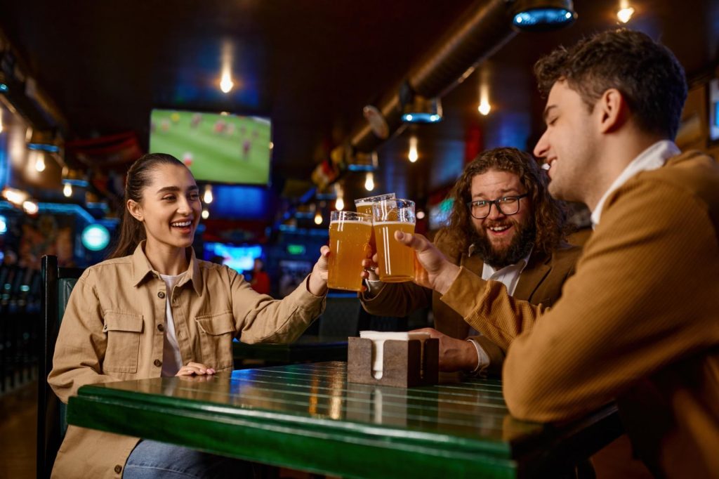 Friends watching football match clinking beer mug in sports bar.