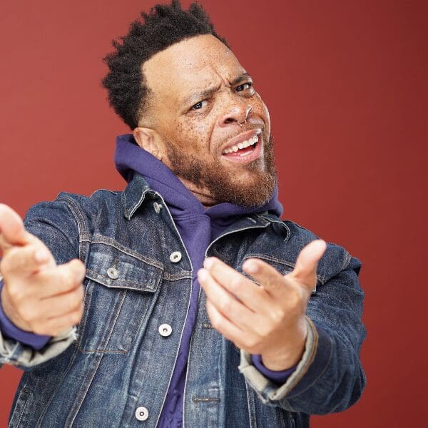 Comedian Tahir Moore in a denim jacket gesturing with a skeptical expression against a red background