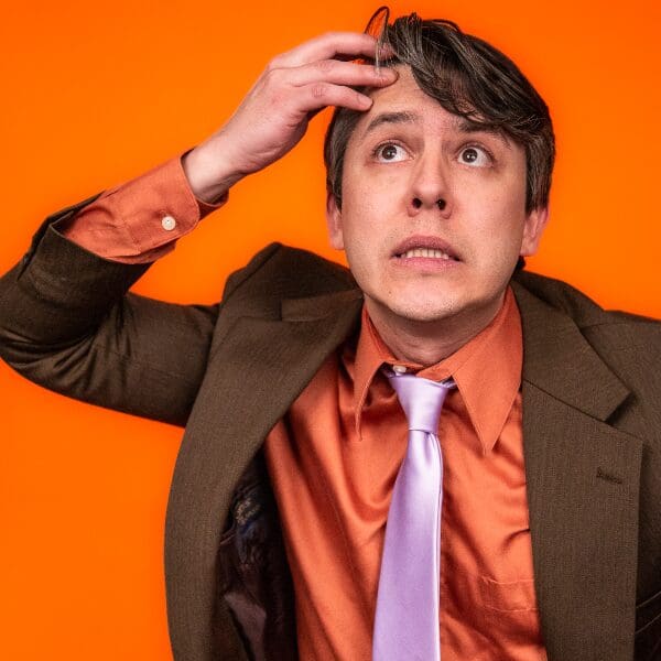 data-30 Comedian Nick Guerra looking up nervously with a hand on his head against an orange background