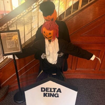 The Delta King scarecrow with a jack-o'-lantern head in a black tailcoat beside a Delta King sign
